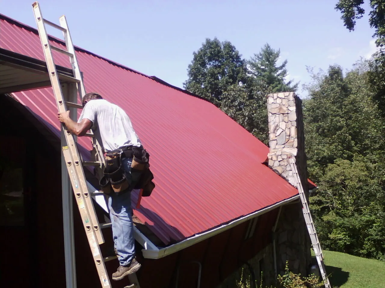 Roofer on a ladder beside a red metal roof and stone chimney — Soffit & Fascia Repair by Roofing Brothers in South Glens Falls